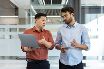 Two professionals are engaged in a discussion, using a laptop and tablet in a modern office setting. Their interaction suggests a collaborative business environment with the use of technology.