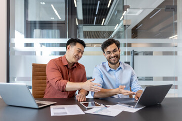 Two male colleagues are engaging in a discussion while working on laptops in contemporary office setting. Papers and smartphone are also visible on the desk, highlighting a collaborative environment.
