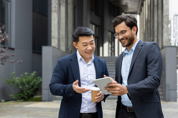 Two professional men in suits having a discussion over a tablet outdoors. One is holding a coffee cup, and both are smiling, signifying a positive and collaborative business interaction.