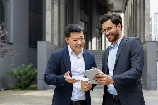Two professional men in suits having a discussion over a tablet outdoors. One is holding a coffee cup, and both are smiling, signifying a positive and collaborative business interaction.