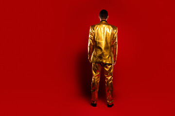 A young man in a shiny golden suit stands with his back facing a vivid red background, exuding elegance and charm