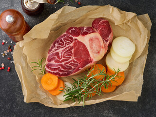Raw osso buco. Fresh veal cut meat on parchment paper with vegetables and spices on an old concrete table background. Italian cuisine background with ingredients.