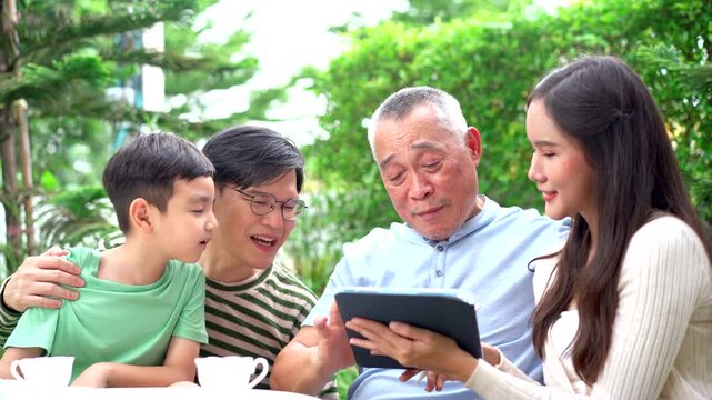 Happy Asian family are sitting in backyard garden at home and talking together. Woman is purchasing a health insurance for her father and let  him review the health insurance packages on tablet.