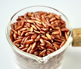 Uncooked raw brown red rice or beras merah food ingredients in small glassware cup pot container isolated on square white background.