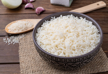 Cooked white rice in a bowl over wooden table with onion, bay leaf and garlic
