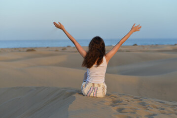 Back view of a girl seating on a sand with hands up at the background of a sea at a golden hour before sunset. Dune in Maspalomas, Gran Canaria. Touristic travel concept