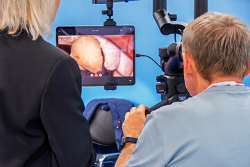 A man looks into the binoculars of a dental microscope. In front of him is a monitor with an image of a jaw and a tooth.