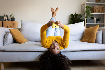 Joyful African American woman lying upside down on the sofa using smart phone. © Daniel