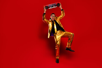 Fototapeta premium A young, handsome man in a glossy gold suit enjoys the moment by dancing energetically with a boombox against a vibrant red background.