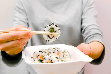 Woman eating fish sushi rolls with salmon,sesame in plastic disposable container box.Seafood,Food delivery,take out service from restaurant at home concept.