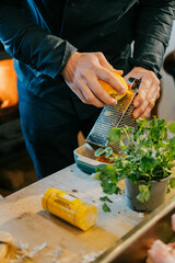 A man is preparing food on a table with a grater and a bottle of mustard. The table is cluttered...