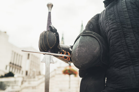 Long medieval sharp sword and protective mask close-up and warrior dressed in black historical fencing armor. Historical items and old traditions and battle restorations, sporty people concept. - Powered by Adobe