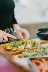 A woman is cutting up vegetables on a cutting board. The vegetables include broccoli and carrots. The woman is wearing a black shirt and jeans
