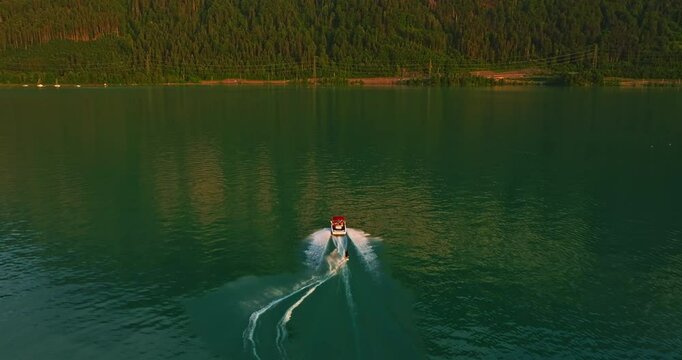 Water skiing dude pulled by the speedboat in the outdoors at blue lake with many luscious, green trees in the background and a cloudy sky, you can see splashes of water or waves. Switzerland