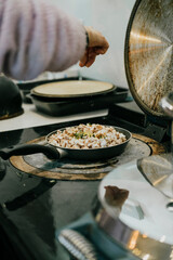 A person sprinkling salt on food in a pan. The pan is on a stove. The person is wearing a pink sweater