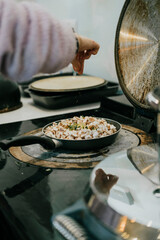 A person sprinkling salt on food in a pan. The pan is on a stove. The person is wearing a pink sweater