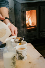 A person is pouring milk into a cup while another person is pouring coffee into a cup. The scene is set in front of a fireplace, with a person pouring coffee into a cup next to a bottle of milk