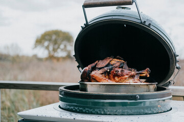 A large piece of meat is cooking on a grill. The grill is open, and the meat is surrounded by a silver pan. The scene is outdoors, and the weather appears to be cloudy