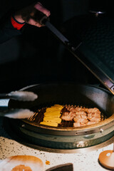 A person is cooking food on a grill. The food includes chicken and yellow items. The person is using tongs to flip the food