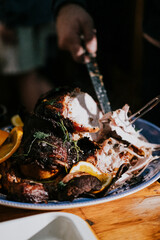 A person is cutting into a large piece of meat on a blue plate. The meat is surrounded by vegetables, including carrots and oranges. The plate is placed on a wooden table
