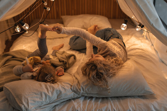 Woman lying in an illuminated play tent playing with her daughter and pulling her foot