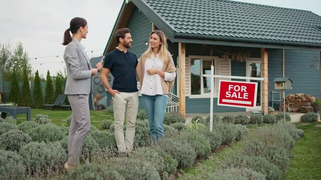 View of Caucasian couple talking with real estate agent. Small building located in background. Red sign with written "For Sale" text. Wife asking questions about property information to realtor.