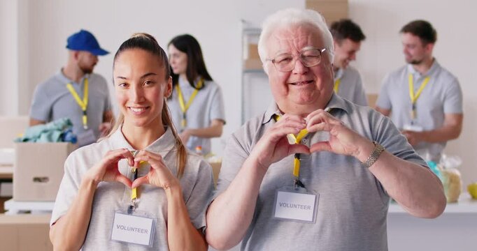 Happy cheerful positive young and senior volunteers doing symbolic heart shape hand sign gesture, smiling and sending you love, support and kind positive feelings. Teamwork in charitable foundation.