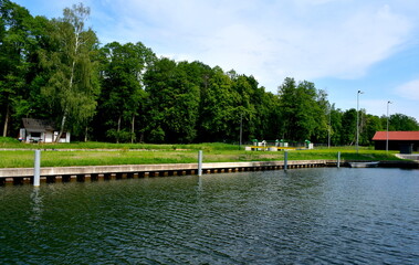 Obraz premium A view of a vast yet shallow river, lake or channel surrounded from all sides with reeds, shrubs and forests with some stone and concrete barriers seen, a small jetty, some boats and a quaint town