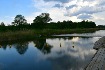 A view of a vast yet shallow river, lake or channel surrounded from all sides with reeds, shrubs and forests with some stone and concrete barriers seen, a small jetty, some boats and a quaint town