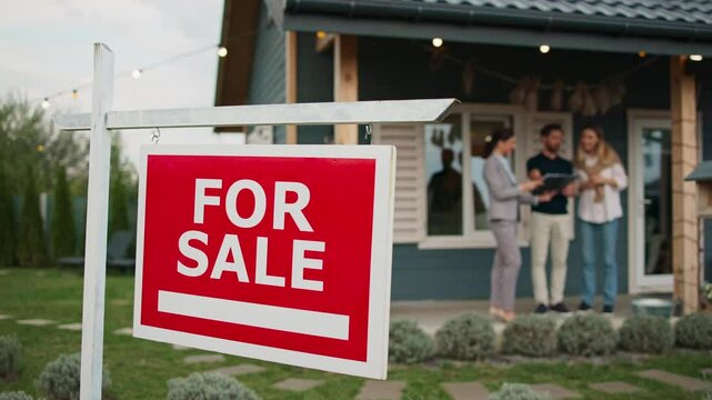 Camera focus on sign hanging in front of entrance to house. In blurred background people discussing with real estate agent possible price for property. Family looking for new home.