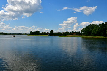 A view of a vast yet shallow river, lake or channel surrounded from all sides with reeds, shrubs and forests with some stone and concrete barriers seen, a small jetty, some boats and a quaint town