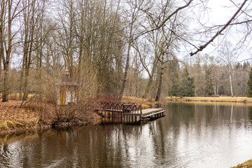 Fototapeta premium Little wooden house and small wooden pier over a calm lake, surrounded by autumn trees with bare branches. The water reflects the sky and foliage, creating 