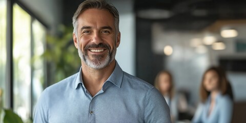 Fototapeta premium A man with a big smile on his face is standing in front of a group of people. He is wearing a blue shirt and a gray beard. The scene appears to be a professional setting