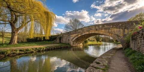 Fototapeta premium A stone arch bridge spans a tranquil canal, surrounded by lush greenery and a weeping willow tree with its branches cascading towards the water, under a blue sky adorned with fluffy white clouds.