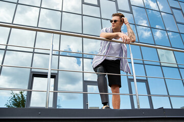 Young man with a prosthetic leg stands stylishly outside a contemporary building under blue skies.