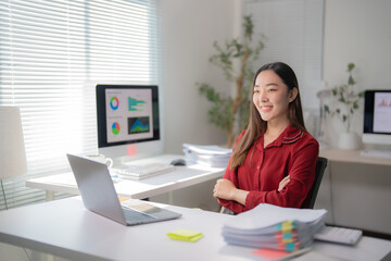 Young asian businesswoman sitting at desk with arms crossed, smiling and looking away while working with laptop and desktop computer showing charts and graphs in modern office