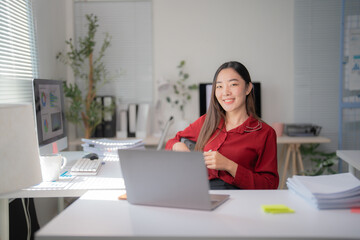 Young asian businesswoman sitting at her desk working with laptop and smiling in modern office, she is wearing a red shirt and is surrounded by paperwork and technology