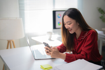 Cheerful young asian office worker sitting at desk using smartphone and laptop while working on project in modern office, happy businesswoman enjoying online communication on mobile phone