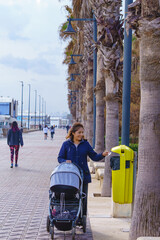 A woman with a baby stroller recycling civically, throws a bag of garbage into a yellow vertical garbage container placed on a city street. walk with palm trees.