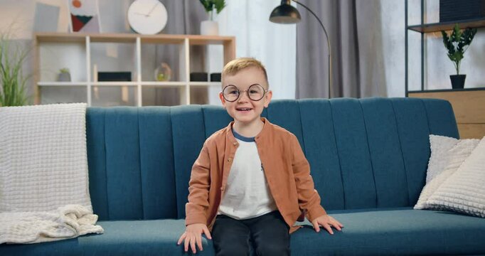Happy boy in glasses dressed in stylish clothes looking into camera with satisfied face sitting on the comfortable couch