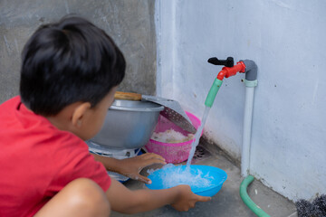 Naklejka premium An Indonesian boy in a red shirt is enthusiastically helping his mother wash the dishes in the sink.