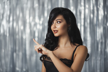Obraz premium Beautiful brunette woman wearing black lingerie and pointing to the side with a sultry expression, set against a glittering silver backdrop. Perfect for luxury, fashion, and promo themes. 