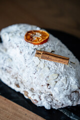Close-up of a classic Christmas Stollen covered in powdered sugar, garnished with dried orange, cinnamon stick on a black plate. Traditional German or Austrian Christmas pastries. Festive atmosphere
