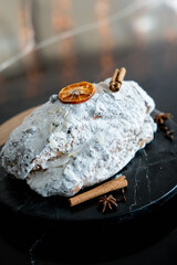 Close-up of a classic Christmas Stollen covered in powdered sugar, garnished with dried orange, cinnamon stick on a black plate. Traditional German or Austrian Christmas pastries. Festive atmosphere