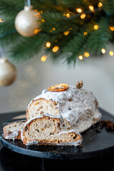 A festive Christmas Stollen covered in powdered sugar, garnished with dried orange and cinnamon, showing sliced portions revealing dried fruit and nuts. Cozy holiday atmosphere  .