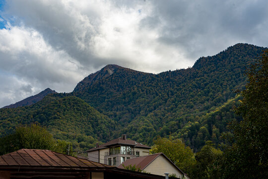 Picturesque mountain resort landscape with low clouds on mountain peaks and low houses in foothill valley on cloudy autumn day. Mountain resort in early autumn - Powered by Adobe