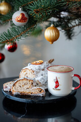 Cup of coffee and  festive Christmas Stollen covered in powdered sugar, garnished with dried orange and cinnamon, showing sliced portions revealing dried fruit and nuts. Cozy holiday atmosphere