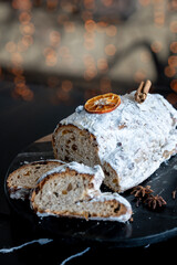 A festive Christmas Stollen covered in powdered sugar, garnished with dried orange and cinnamon, showing sliced portions revealing dried fruit and nuts. Cozy holiday atmosphere  .