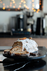 A festive Christmas Stollen covered in powdered sugar, garnished with dried orange and cinnamon, showing sliced portions revealing dried fruit and nuts. Cozy holiday atmosphere  .