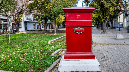 A vibrant red British-style postbox stands on a tree-lined city street, symbolizing communication and nostalgia